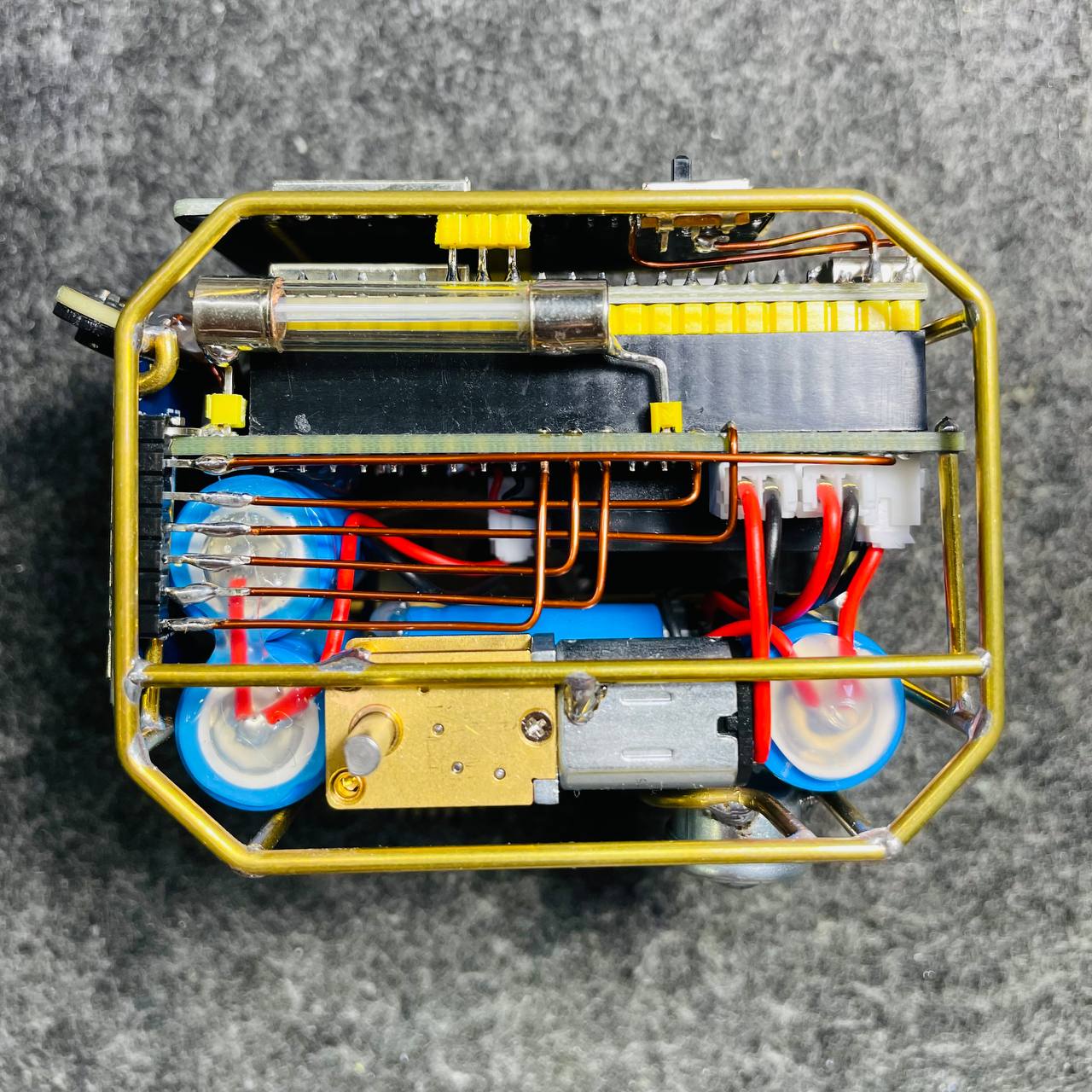 Bên trong VuiTV Robot V2 nhìn từ trên xuống với khung vàng đồng, bo mạch, dây nguồn, động cơ, hộp giảm tốc và hai bánh xe xanh